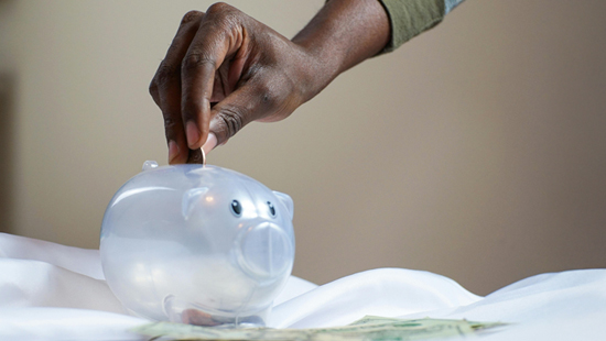 a person of color with a green sleeve inserts a coin into a clear piggy bank